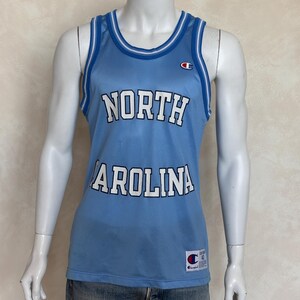 May include: A light blue mesh basketball jersey featuring the text NORTH CAROLINA in white letters with black outlines. The sleeveless top has white and blue striped trim on the neck and arm openings, a Champion logo on the chest, and a label at the hem.