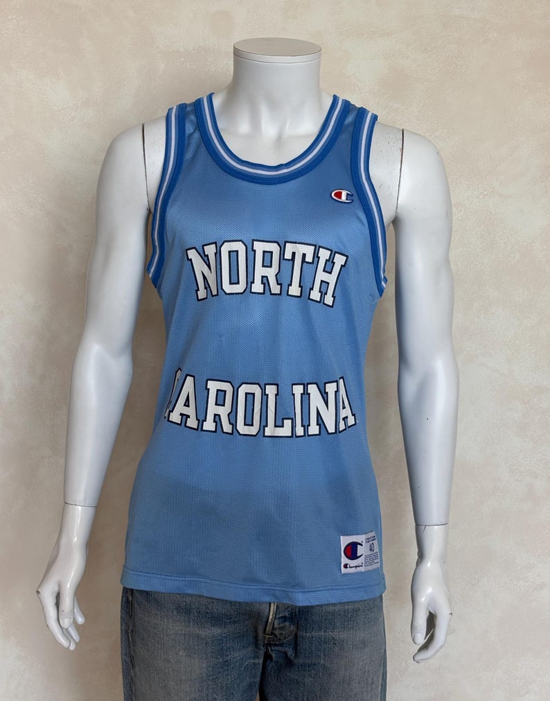 May include: A light blue mesh basketball jersey featuring the text NORTH CAROLINA in white letters with black outlines. The sleeveless top has white and blue striped trim on the neck and arm openings, a Champion logo on the chest, and a label at the hem.