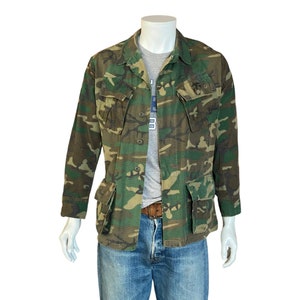Puede incluir: Chaqueta de camuflaje con cuatro bolsillos delanteros y cierre de botones. La chaqueta, en tonos verdes, marrones y beige, se exhibe en un maniquí. Tiene cuello y mangas largas.