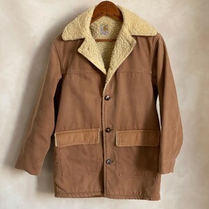 May include: A brown corduroy jacket with a sherpa lining and a Carhartt logo on the pocket. The jacket has a button closure and two large pockets on the front.