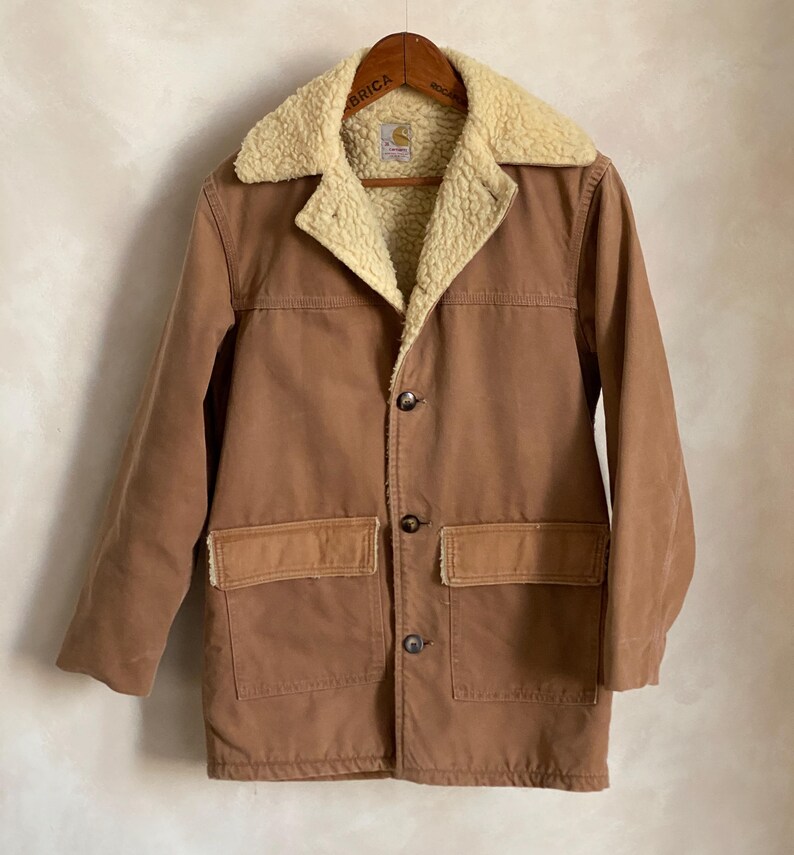 May include: A brown corduroy jacket with a sherpa lining and a Carhartt logo on the pocket. The jacket has a button closure and two large pockets on the front.