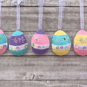 May include: Five felt Easter egg ornaments hanging from white ribbon. The eggs are decorated with pastel colors, embroidered flowers, and sequins.