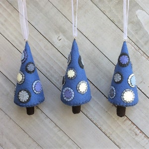 May include: Three blue felt Christmas tree ornaments with white, gray, and blue polka dots. The ornaments are hanging from white ribbon.