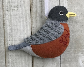 May include: A felt robin ornament with a gray back, orange breast, and black head. The robin has a yellow beak and black eyes. The ornament is hanging from a white ribbon.