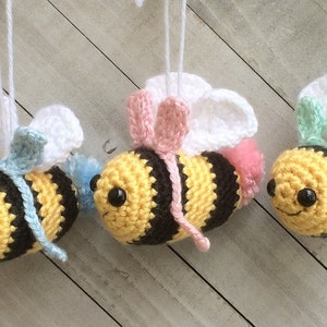 May include: Three crocheted bee ornaments with white wings, each with a different colored bow: blue, pink, and green. The bees are hanging from white strings.