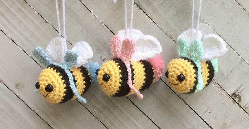 May include: Three crocheted bee ornaments with white wings, each with a different colored bow: blue, pink, and green. The bees are hanging from white strings.