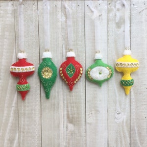 May include: Five felt Christmas ornaments hanging from white ribbon. The ornaments are red, green, yellow, and white with gold glitter and sequins.