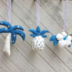 May include: Three felt ornaments in the shape of a palm tree, pineapple, and a flower. The ornaments are white with blue accents and are decorated with sequins.