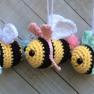 May include: Three crocheted bee ornaments with white wings, yellow and black striped bodies, and different colored accents. The bees are hanging from white strings.