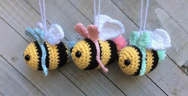 May include: Three crocheted bee ornaments with white wings, yellow and black striped bodies, and different colored accents. The bees are hanging from white strings.