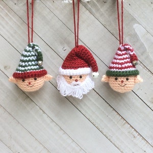 May include: Three crocheted Christmas ornaments hanging from red strings. The ornaments are a Santa Claus, an elf, and another elf. The Santa Claus ornament has a red hat, white beard, and black eyes. The elves have green and white striped hats, red and white striped hats, and black eyes.