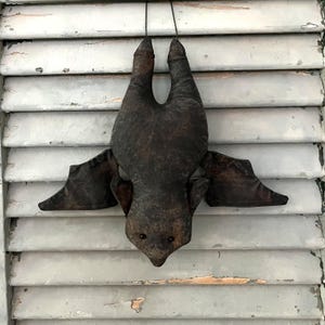 May include: A dark brown bat-shaped plush decoration with outstretched wings, hanging from a black string. The bat has small, round eyes and a textured, aged appearance, suggesting a vintage or rustic style. The wings are a darker shade of brown.