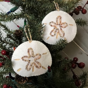 May include: Two white felt sand dollar ornaments with beaded star designs hanging on a Christmas tree.