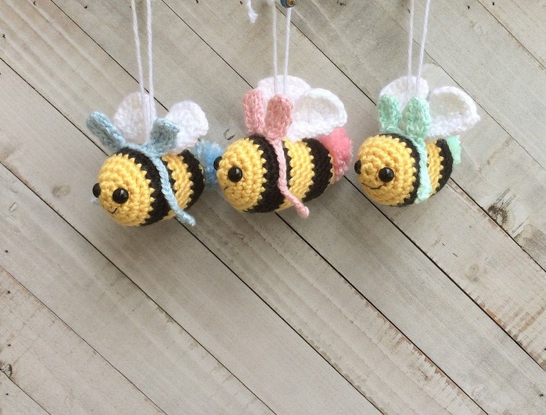 May include: Three crocheted bee ornaments with white wings, black and yellow striped bodies, and different colored bows. The bees are hanging from white strings against a light wood background.