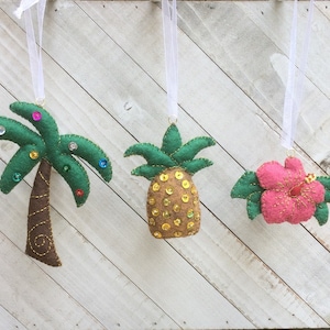 May include: Three felt ornaments, a green palm tree, a brown pineapple, and a pink hibiscus flower. Each ornament has a white ribbon for hanging.