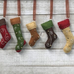 May include: Five felt Christmas stockings with different colored felt and embroidered designs. Each stocking has a brown ribbon loop for hanging.