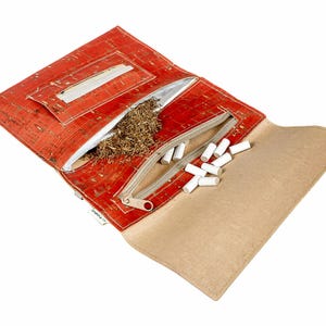 Può includere: Una custodia per tabacco in sughero rosso, aperta per mostrare tabacco e filtri bianchi. La custodia ha uno scomparto con cerniera e una sezione separata. Lo sfondo è bianco.