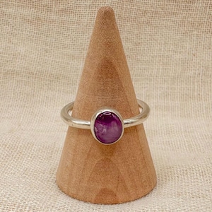 May include: A silver ring with a purple oval gemstone set in the centre. The ring is displayed on a wooden ring holder.