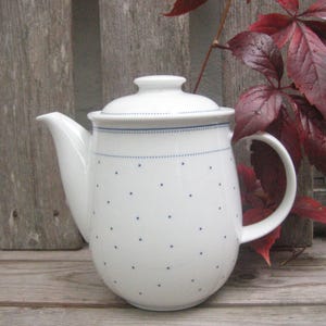 May include: White ceramic teapot with a lid, decorated with small blue polka dots and a blue dotted line. The teapot has a curved spout and handle. The background features wooden planks and red leaves.