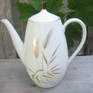 Peut inclure: Une cafetière en porcelaine blanche avec un corps effilé, une anse incurvée et un long bec verseur. L'extérieur est orné d'une illustration délicate de tiges de blé dans des tons dorés, bruns et verts. Elle comprend un couvercle avec un bouton doré.