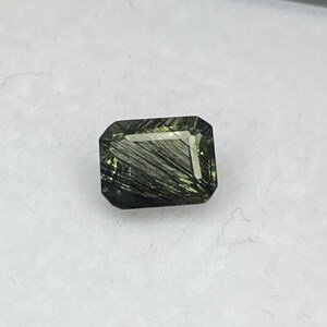 May include: A rectangular-cut, dark green gemstone with internal needle-like inclusions. The gem is set against a white background, showcasing its clarity and colour. The stone's facets reflect light, highlighting its polished surface.