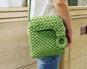 May include: A green crocheted crossbody bag with a round detail on the front flap. The bag is worn over the shoulder of a person wearing a white shirt and blue jeans.