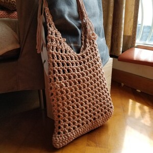 May include: A light brown crocheted tote bag with a wide, open top and long fringe handles.