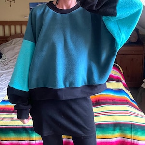 Może przedstawiać: Bluza z polaru z długim rękawem o wzorze typu color-block w kolorach turkusowym, jasnym cyjanowym i czarnym. Odzież posiada okrągły dekolt, czarne ściągacze przy rękawach oraz czarny dół. Główna część jest turkusowa z cyjanowymi i czarnymi wstawkami.