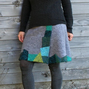 Könnte beinhalten: Eine Person trägt einen dunkelgrauen Pullover, einen grauen Rock mit grünen und blauen Patchwork-Elementen und gestreifte Leggings. Sie trägt auch einen grünen Schal und eine Sonnenbrille. Der Hintergrund ist eine verwitterte Holzwand.