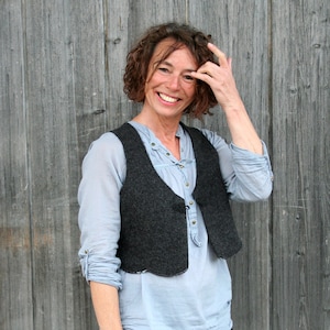 Peut inclure: Une femme aux cheveux bruns bouclés porte une chemise bleu clair avec un gilet gris foncé et un pantalon noir. Elle sourit et a la main sur le visage.
