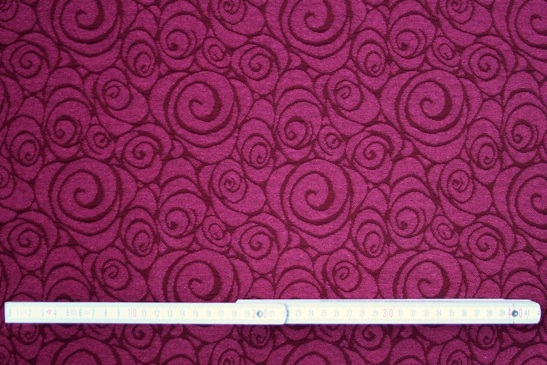Peut inclure: Gros plan sur un tissu avec un motif de tourbillons r&eacute;p&eacute;t&eacute;s dans des tons bordeaux et marron fonc&eacute;. Une r&egrave;gle gradu&eacute;e en centim&egrave;tres est plac&eacute;e en bas, indiquant l'&eacute;chelle du motif. Le tissu pr&eacute;sente un aspect textur&eacute;.