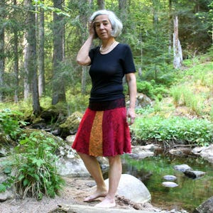 Könnte beinhalten: Eine Person trägt ein schwarzes Oberteil mit kurzen Ärmeln und einen knielangen Rock mit roten, orangefarbenen und goldenen vertikalen Bahnen. Der Rock hat einen Spitzensaum an der Taille. Die Person steht in einem Wald in der Nähe eines Baches.