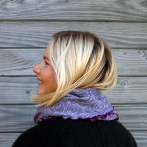 Könnte beinhalten: Eine Frau mit blondem Haar trägt einen lila Schal mit einem weißen Blumenmuster und einem dunkelvioletten, gerüschten Rand. Der Schal liegt um den Hals, und die Frau lächelt. Der Hintergrund ist eine Holzwand.