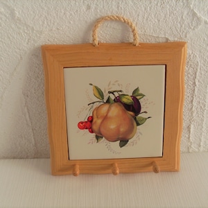 Könnte beinhalten: Eine Holzrahmenfliese mit weißem Hintergrund und einer bunten Illustration von Früchten. Die Fliese hat drei Haken an der Unterseite.