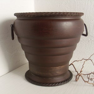 Könnte beinhalten: Ein brauner Metalltopf mit geripptem Design und einer dekorativen Kordelverzierung am oberen und unteren Rand. Der Topf hat zwei Metallgriffe an den Seiten.