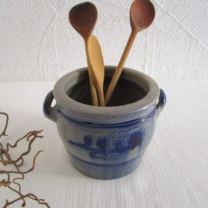 Könnte beinhalten: Ein blau-grauer Keramiktopf mit zwei Griffen, der drei Holzlöffel enthält. Der Topf hat ein blaues Blumendesign und steht vor einem weißen Hintergrund. Die Löffel sind in verschiedenen Brauntönen gehalten.