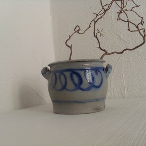 May include: A white ceramic pot with blue painted designs, featuring a dried branch with a few leaves. The pot has a rounded shape and two handles.