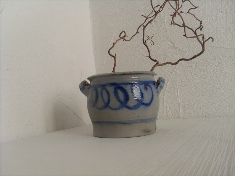 May include: A white ceramic pot with blue painted designs, featuring a dried branch with a few leaves. The pot has a rounded shape and two handles.