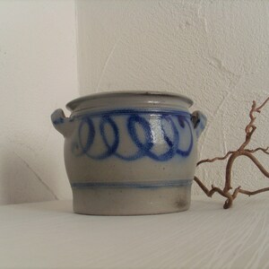 May include: A white ceramic crock with blue painted swirls and two handles. The crock is sitting on a white surface.