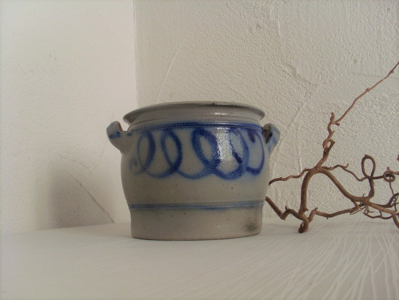 May include: A white ceramic crock with blue painted swirls and two handles. The crock is sitting on a white surface.