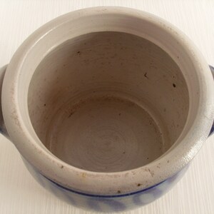 May include: A white ceramic crock with blue and white patterned accents. The crock has two handles and is empty.