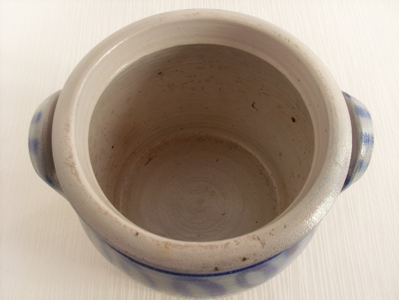 May include: A white ceramic crock with blue and white patterned accents. The crock has two handles and is empty.