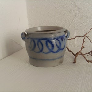 May include: A small, white ceramic pot with blue and gray painted designs. The pot has two handles and a rounded shape.