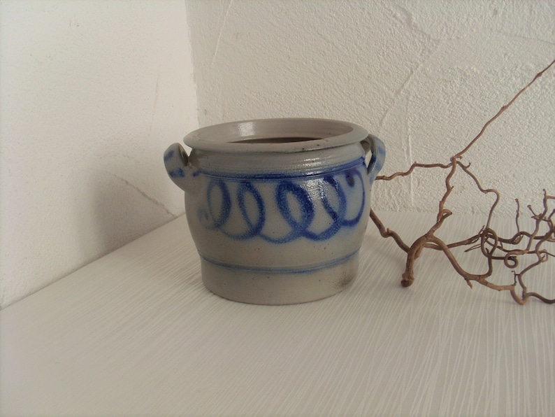 May include: A small, white ceramic pot with blue and gray painted designs. The pot has two handles and a rounded shape.
