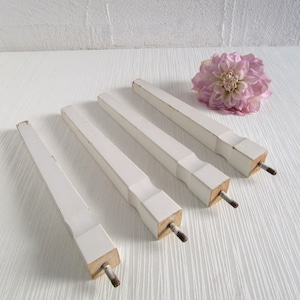 Könnte beinhalten: Vier weiße Holzmöbelfüße mit rechteckigem Design. Jeder Fuß hat eine Metallschraube, die aus der Unterseite herausragt. Im Hintergrund befindet sich eine rosa Blume. Die Beine sind wahrscheinlich für einen Tisch oder andere Möbel.