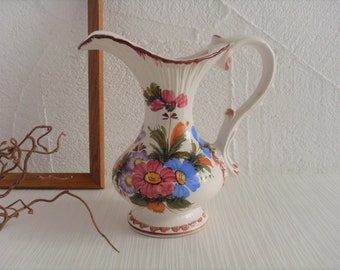 May include: A white ceramic pitcher with a floral design. The pitcher has a wide mouth and a handle. The flowers are painted in shades of pink, blue, orange, and green.