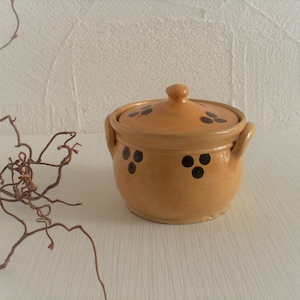 Könnte beinhalten: Ein kleiner, hellbrauner Keramiktopf mit Deckel und schwarzen Punktmustern. Der Topf hat zwei Griffe und einen Knauf auf dem Deckel. Der Hintergrund ist eine strukturierte weiße Oberfläche.