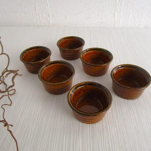 Könnte beinhalten: Ein Set aus sechs kleinen, runden, braunen Keramik-Förmchen mit einer glänzenden, gesprenkelten Glasur. Jede Schale hat einen leicht ausgestellten Rand und ein glattes, gleichmäßiges Inneres, angeordnet auf einer weißen Oberfläche.