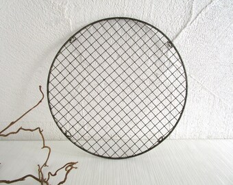 May include: A round, black metal grill grate with a diamond pattern. The grate is set against a white textured wall and a white wooden surface. A few brown twigs are in the lower left corner.