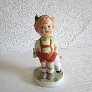 Könnte beinhalten: Eine Porzellanfigur eines Jungen mit roten Haaren und grünen Blättern im Haar, der eine rote Latzhose und ein gelbes Hemd trägt und ein weißes Lamm hält. Die Figur steht auf einem grün-weißen Sockel.